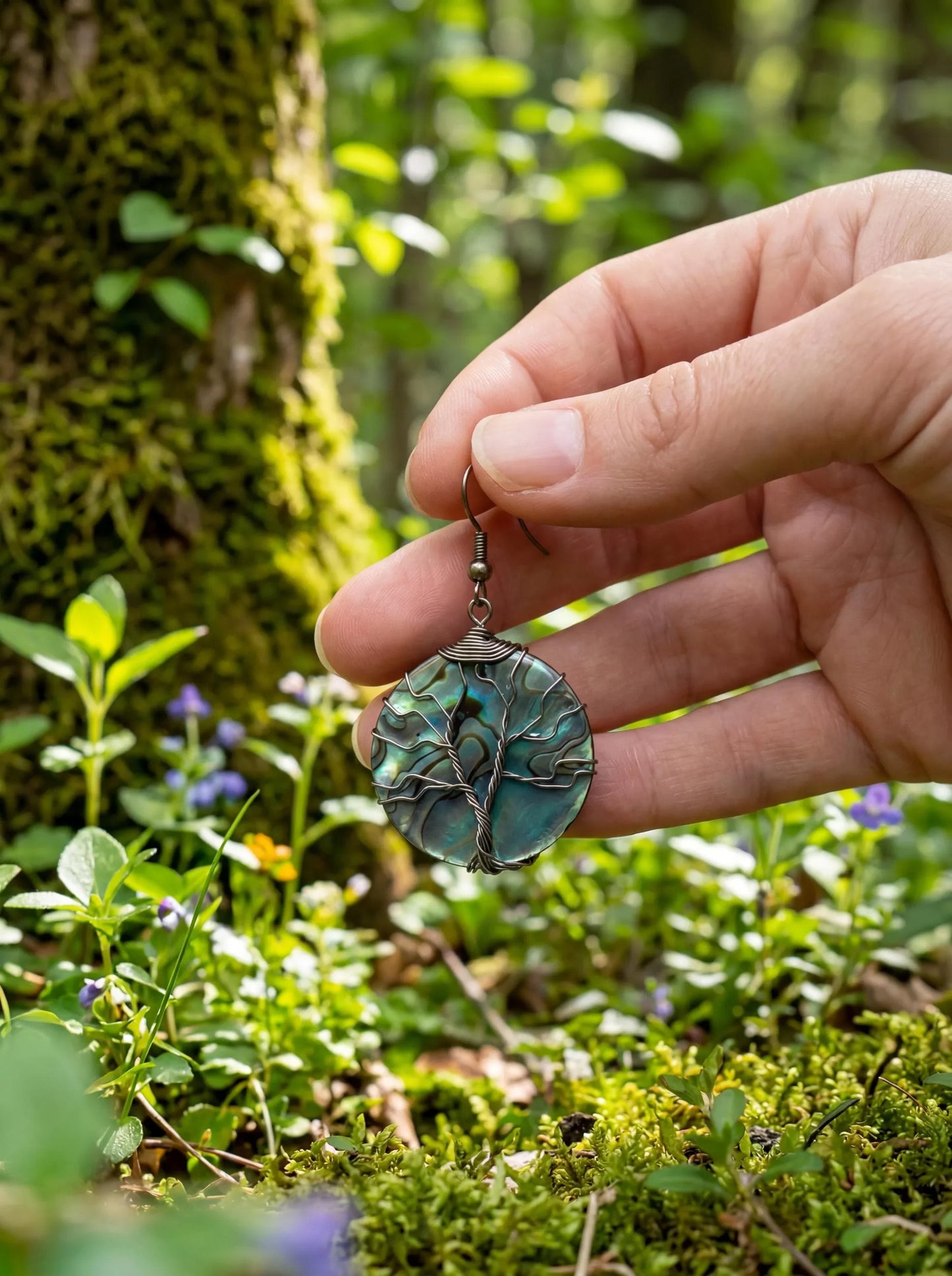 Hand holding wire-wrapped abalone shell earring with tree of life design in forest setting