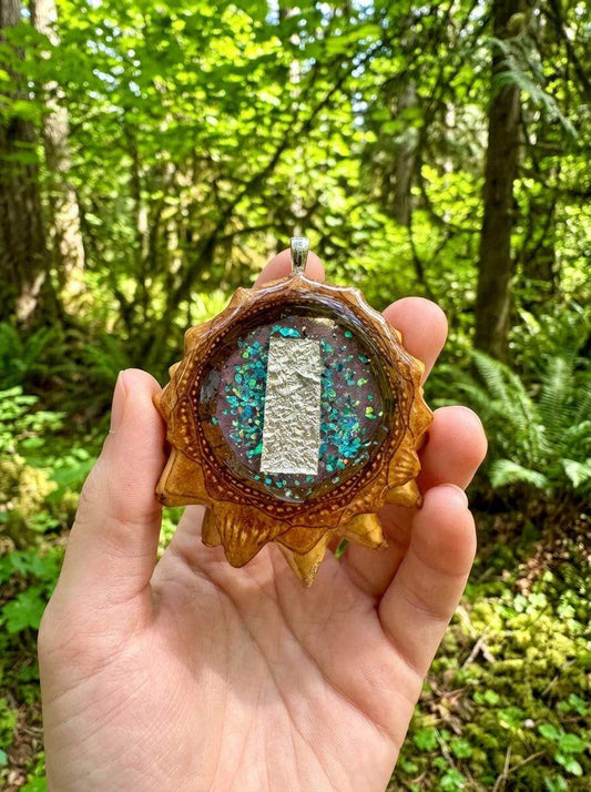Hand holding resin pinecone pendant with gold leaf and blue glitter, forest background