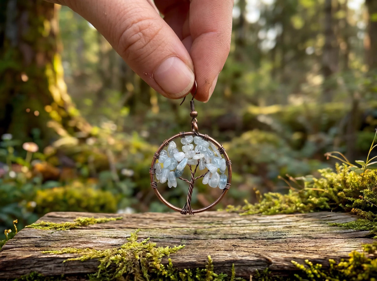 Hand holding wire-wrapped tree of life earring with light blue gemstones, forest moss background