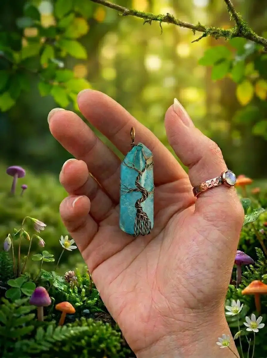 Hand holding blue wire-wrapped pendant with tree of life design, forest backdrop, mushrooms, and wildflowers
