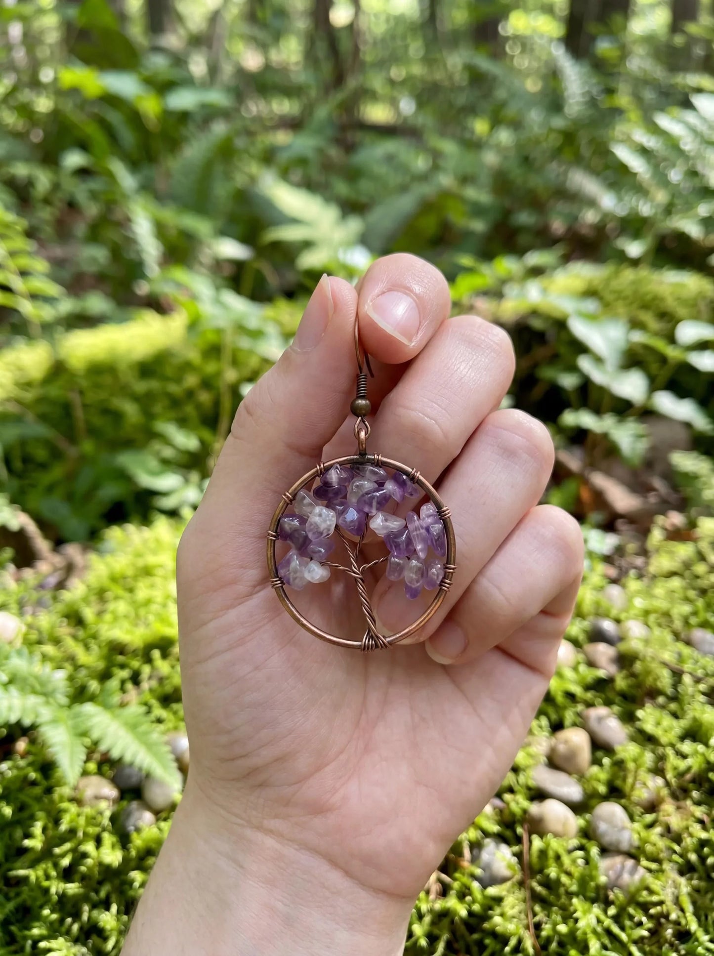 Hand holding copper wire wrapped amethyst tree of life earring in forest setting