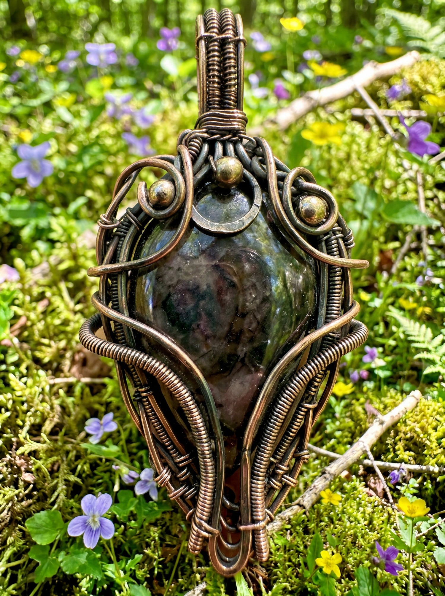 a wire wrapped pendant sitting a top some green moss with a bright and vibrant forest background