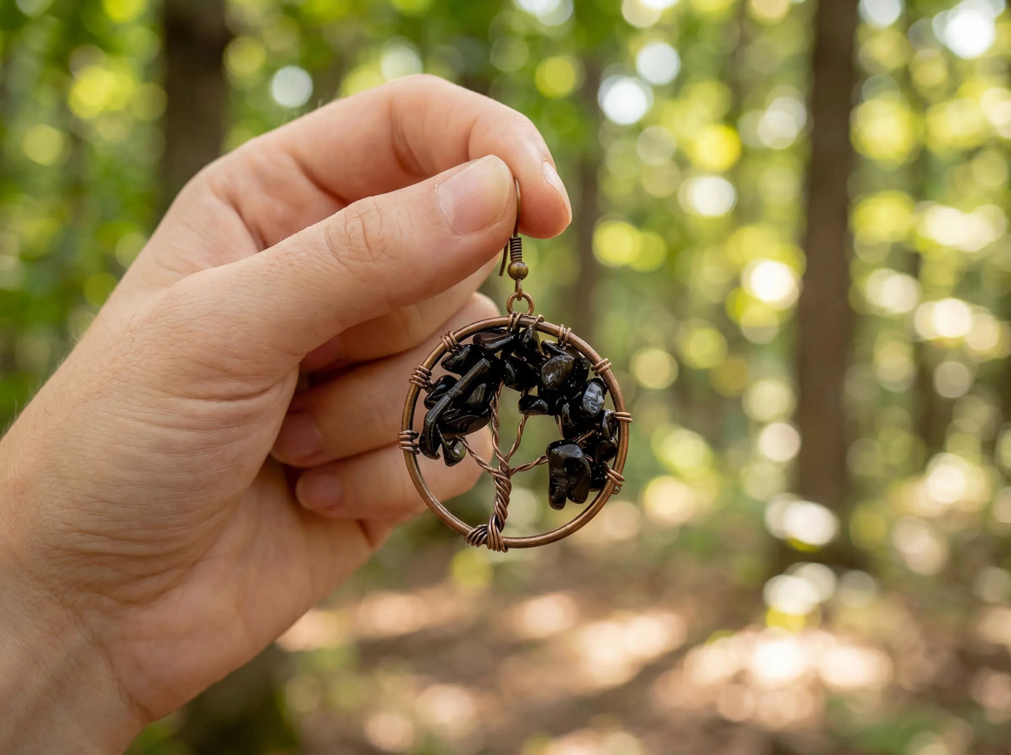 Hand holding copper wire wrapped earring with black gemstones, forest bokeh background