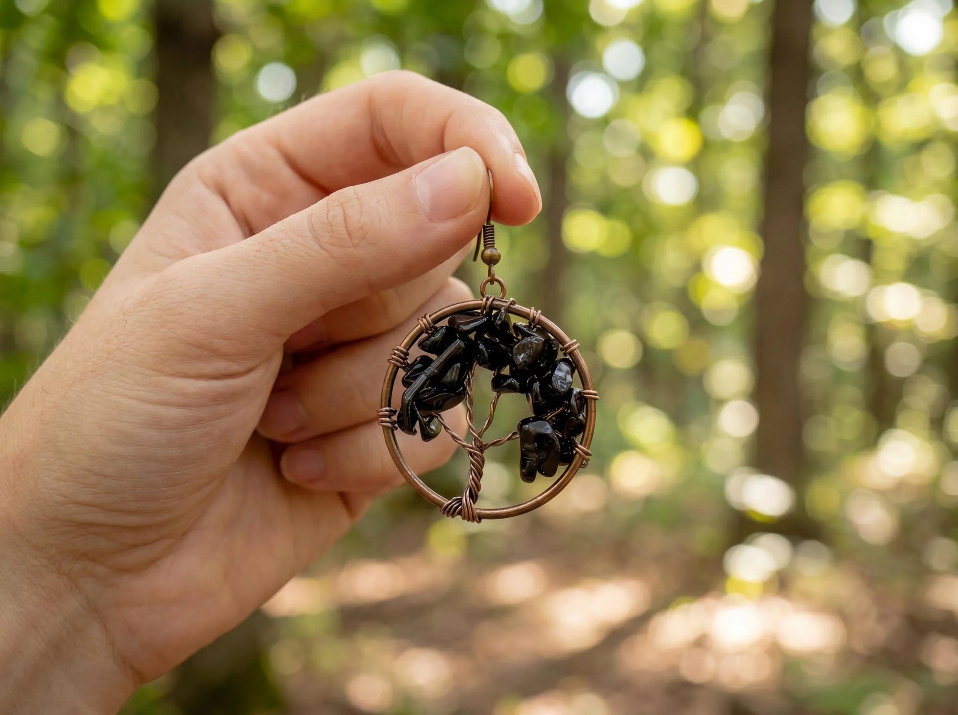 Hand holding copper wire wrapped earring with black gemstones, forest bokeh background