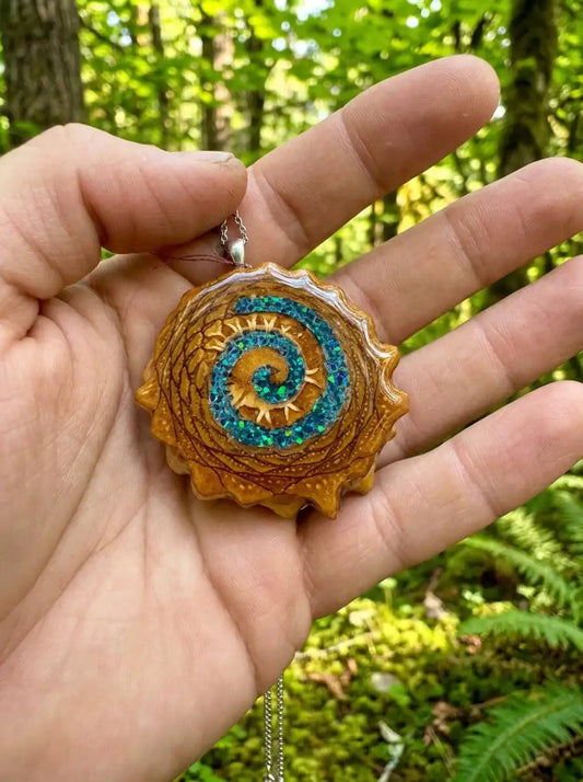 Hand holding resin pinecone pendant with blue spiral glitter, forest background