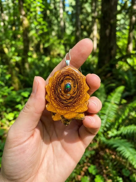 Hand holding resin pinecone pendant with opal center, forest background