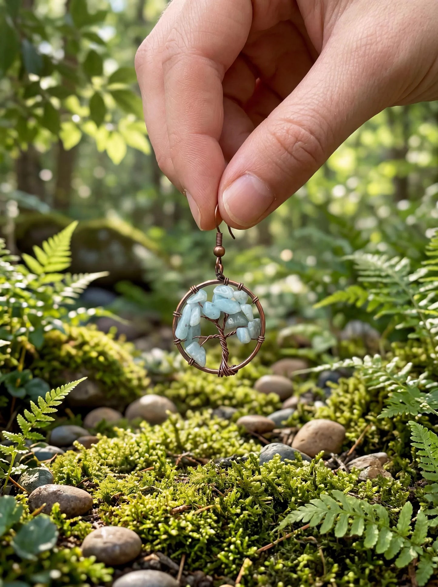 Hand holding wire-wrapped tree of life earring with light blue stones, set against mossy forest floor
