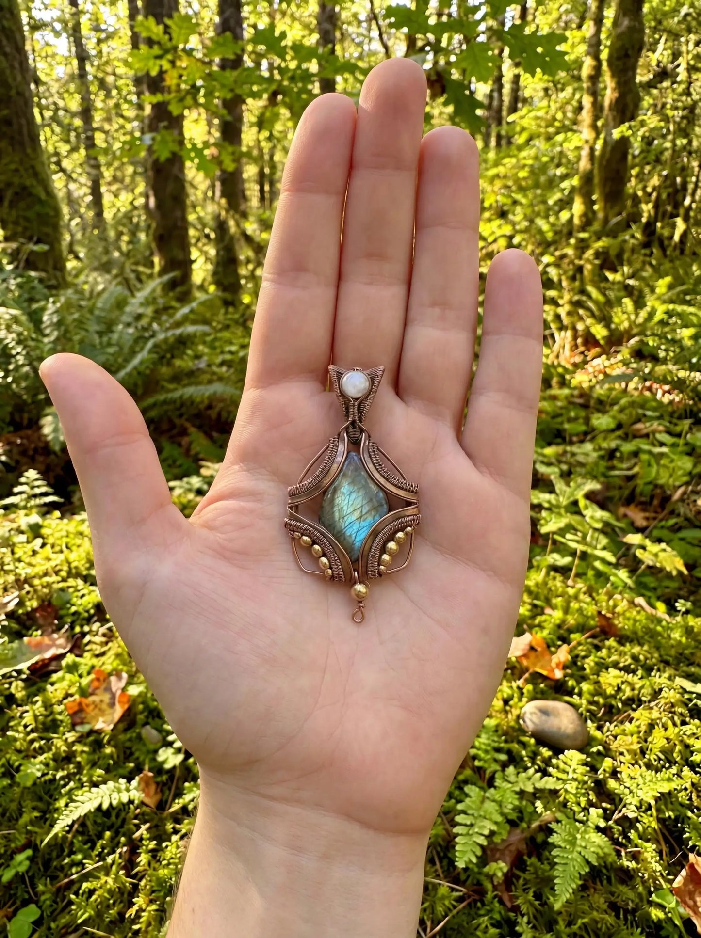 Hand holding wire-wrapped labradorite pendant with moonstone accent in forest setting