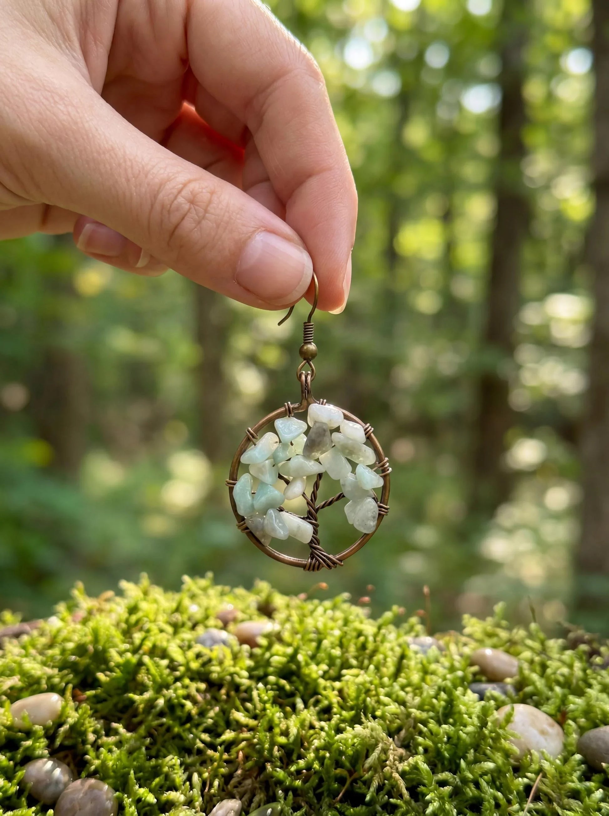 Hand holding wire-wrapped tree of life earring with green gemstones over mossy forest floor
