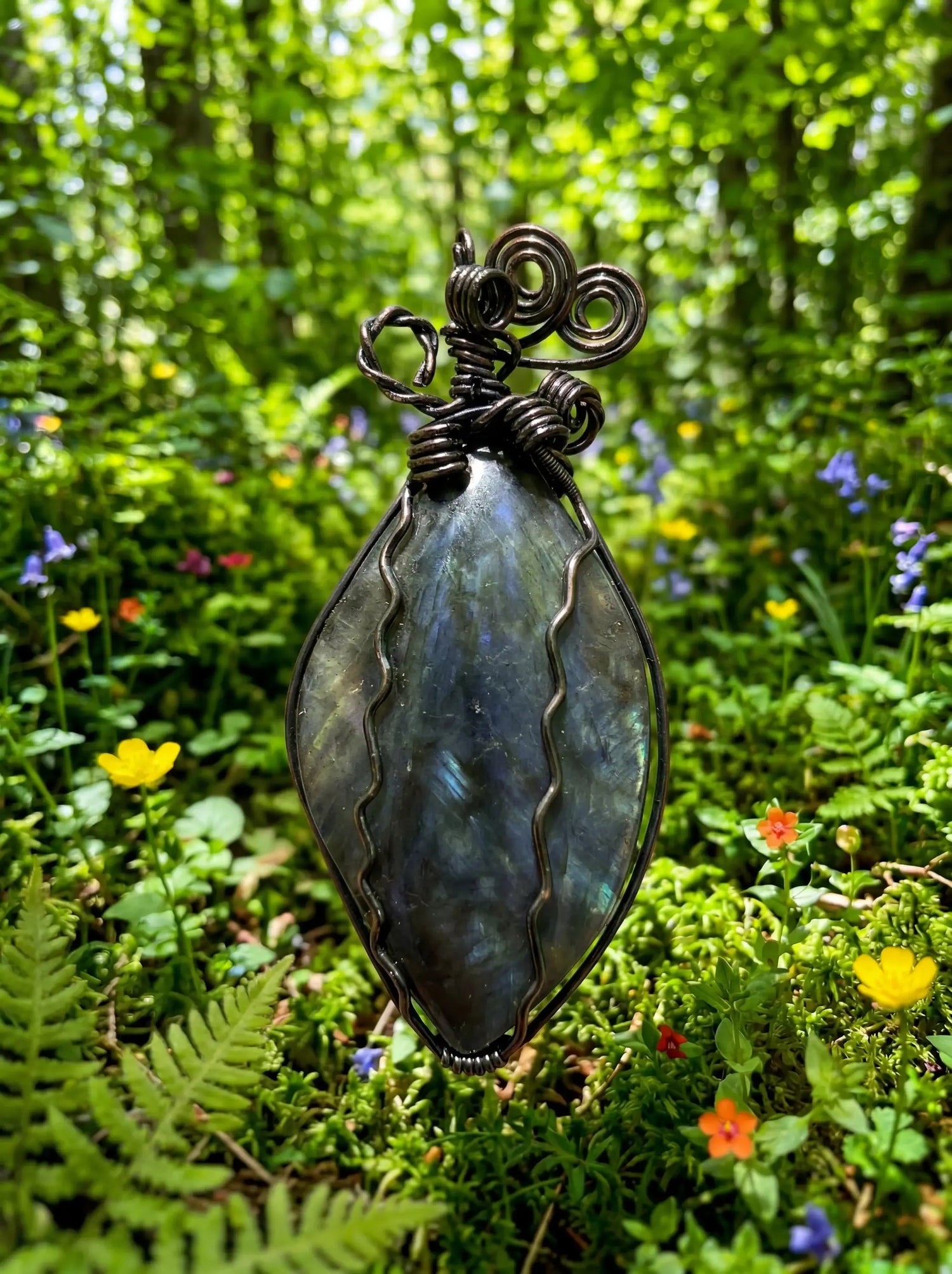 Handcrafted wire-wrapped labradorite pendant with swirling metal design, set against a vibrant forest backdrop.