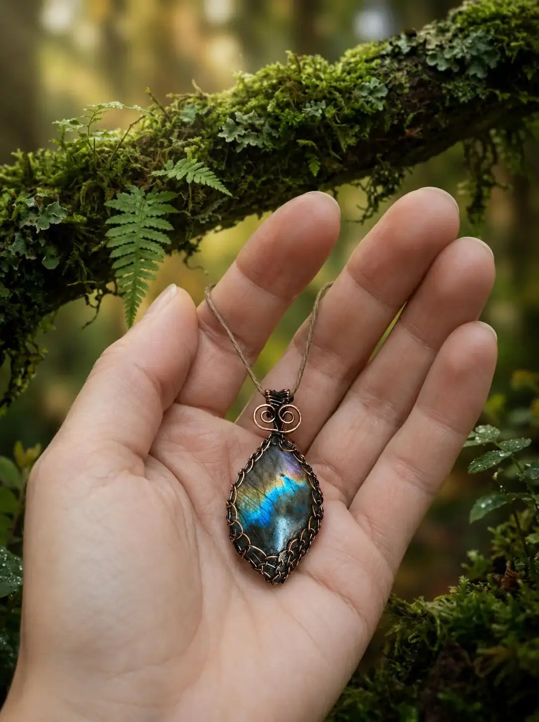 Hand holding wire-wrapped labradorite pendant with mossy forest background, artisan jewelry